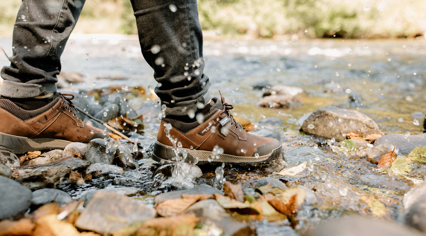 A man wearin a pair of brown nubuck low-top, lace-up splashes in a shallow river stream. The shoes have a rugged black sole, gray mid-sole, waterproof insignia and mesh fabric around the ankle collar. The shoe style is Raymond 51 in color Tabak made by Josef Seibel.